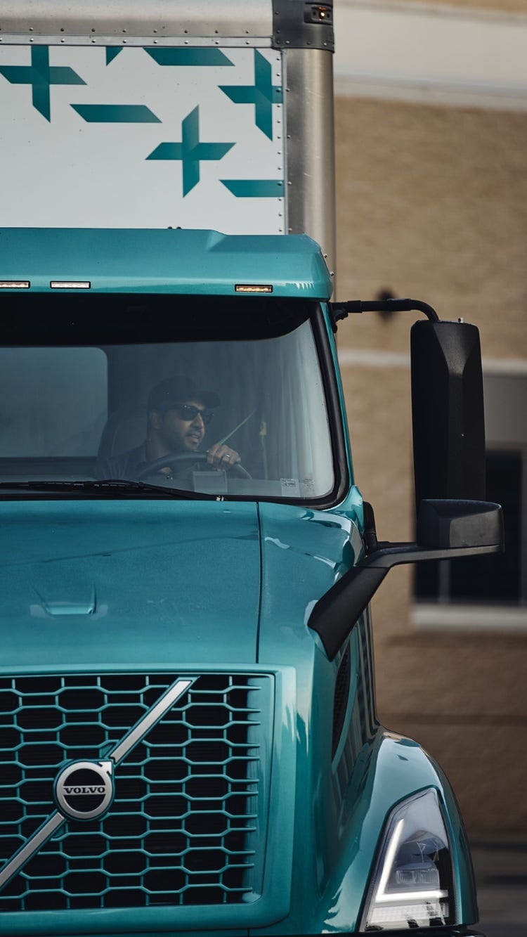 A close-up of the front of a Volvo VNR Electric truck, with a driver visible through the windshield. The truck is teal-colored with modern headlights and a distinctive grille.