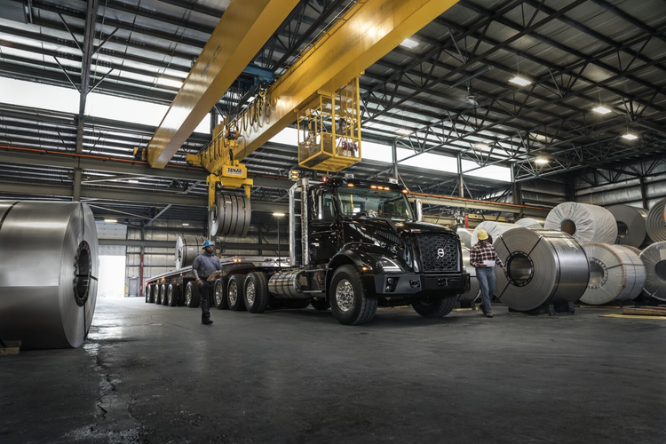A black Volvo VNX truck inside an industrial warehouse, loaded with large steel coils using an overhead crane