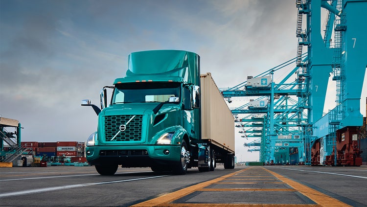 A teal Volvo big truck hauling a shipping container at a port, highlighting sustainable transportation options