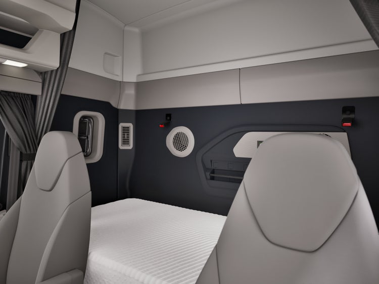 A view of the inside of the Volvo VNX. Two cream colored seats with a white color sleeper space behind the seats.