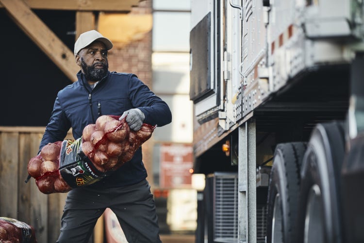 A man in a cap and jacket unloads a large bag of onions from a truck near a loading dock.