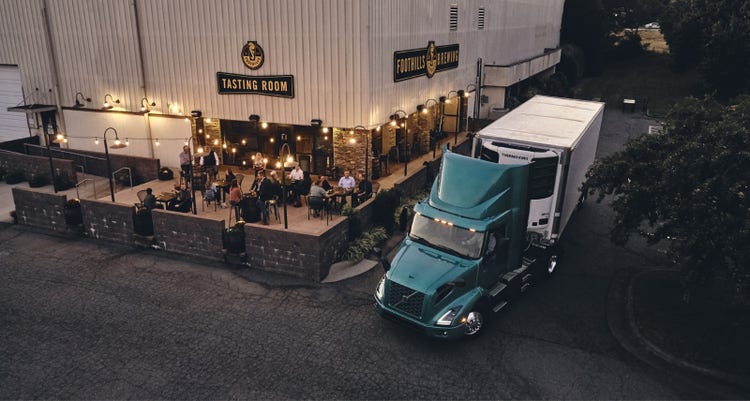 A teal Volvo VNR Electric truck with a box trailer parked outside a brewery's tasting room, where people are seated at outdoor tables under warm lighting.