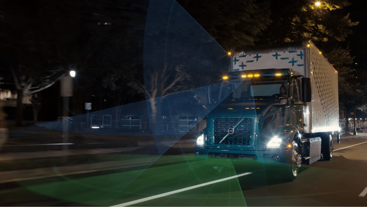 A Volvo VNR Electric truck driving at night on a city street, illuminated by headlights. Overlay graphics represent advanced safety features in action.