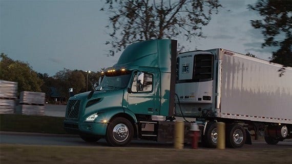 A teal Volvo VNR Electric truck pulling a white trailer, driving past trees and industrial storage during dusk.