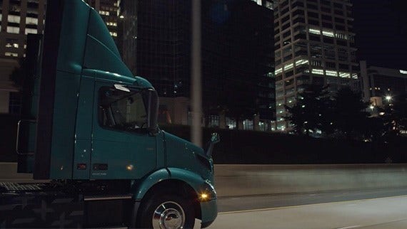 A teal Volvo VNR Electric truck driving through an urban area at night, with illuminated office buildings in the background.
