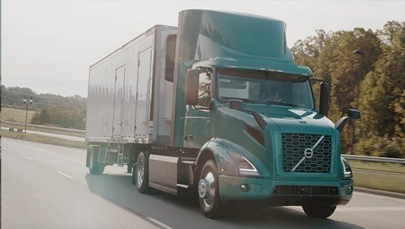 A teal Volvo VNR Electric truck pulling a white trailer, driving on a highway bordered by trees and grassy fields.