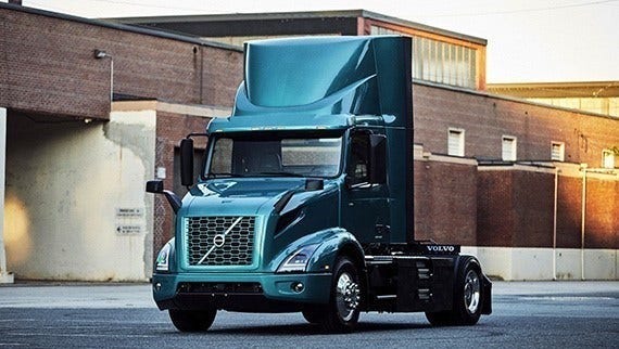 A teal Volvo VNR Electric truck parked in an industrial area, surrounded by brick buildings and a paved lot.