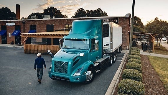 A teal Volvo VNR Electric truck parked outside a brick building with a person walking nearby, surrounded by landscaping and a paved lot.