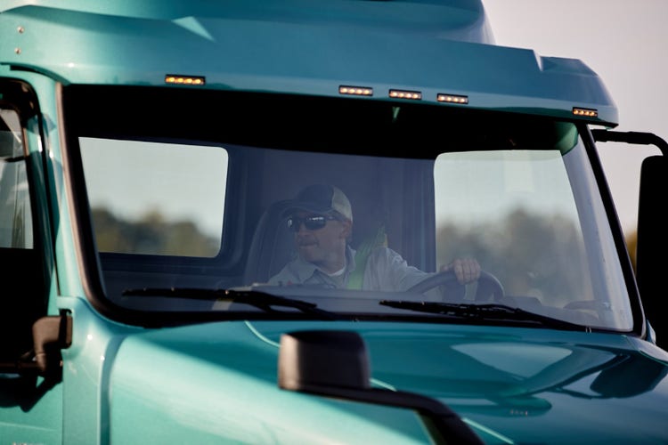Close-up of a driver inside a teal Volvo VNR Electric truck, wearing sunglasses and a cap, visible through the windshield.