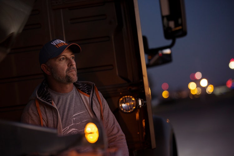 A truck driver wearing a Volvo Trucks cap and hoodie leans against a Volvo VAH truck