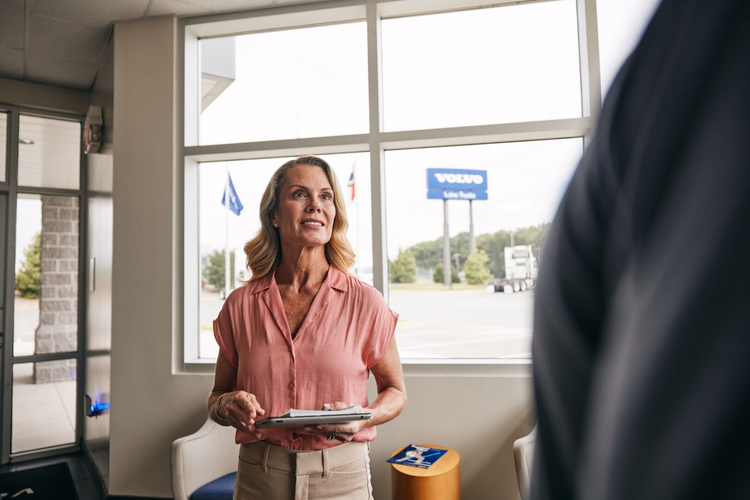 A Volvo representative holding a tablet stands inside a dealership, with a Volvo sign visible through the window in the background.