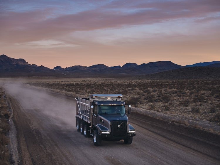 A Volvo VHD truck driving on a dusty desert road at dusk, set against a backdrop of rugged mountains and a colorful evening sky.