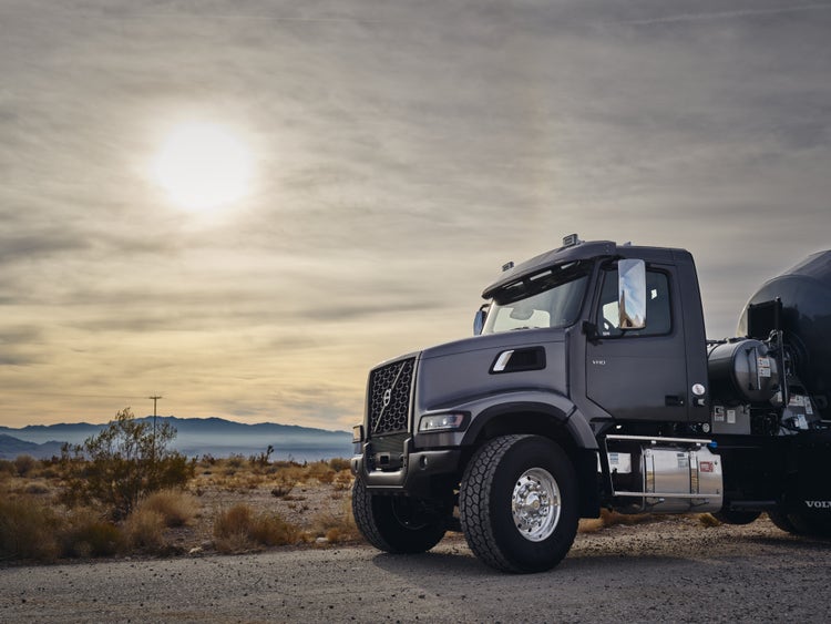 A Volvo VHD truck with a mixer attachment parked in a desert landscape under a bright sun and cloudy sky, with distant mountains in the background.