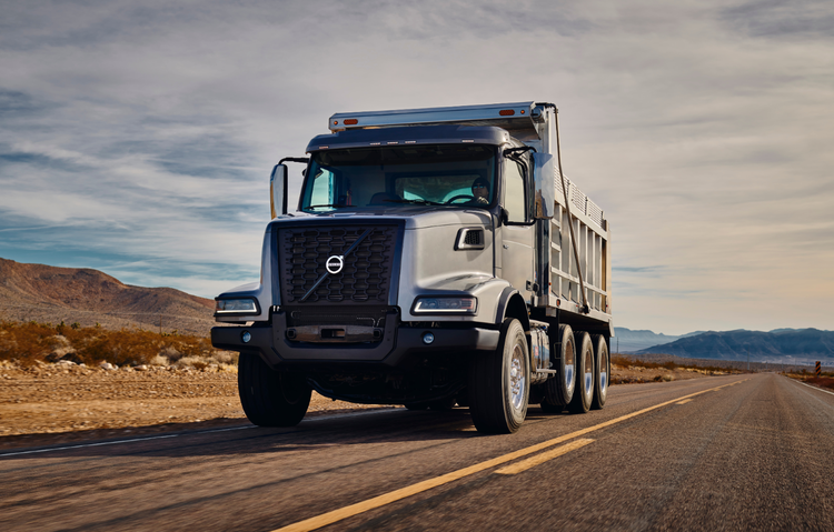A Volvo VHD Axle Back dump truck driving on an open highway surrounded by a desert landscape and distant mountains.