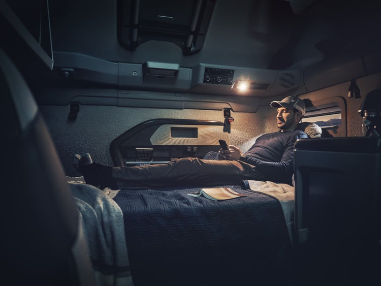 A man relaxes on a bed in a Volvo VNL sleeper cab, holding a phone under a reading light.