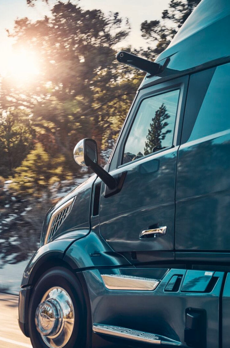 Side view of a Volvo VNL truck cab, highlighting the chrome details and aerodynamic design, with a sunlit forest in the background.