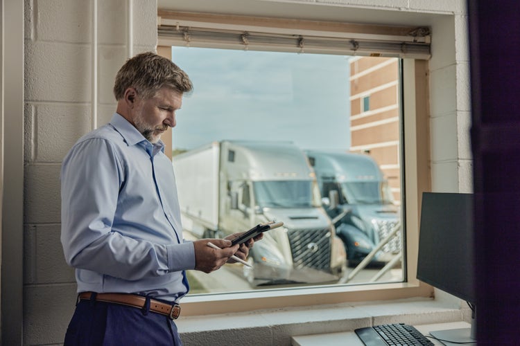 A man in a blue shirt standing near a window, using a tablet, with two Volvo VNL trucks visible outside.