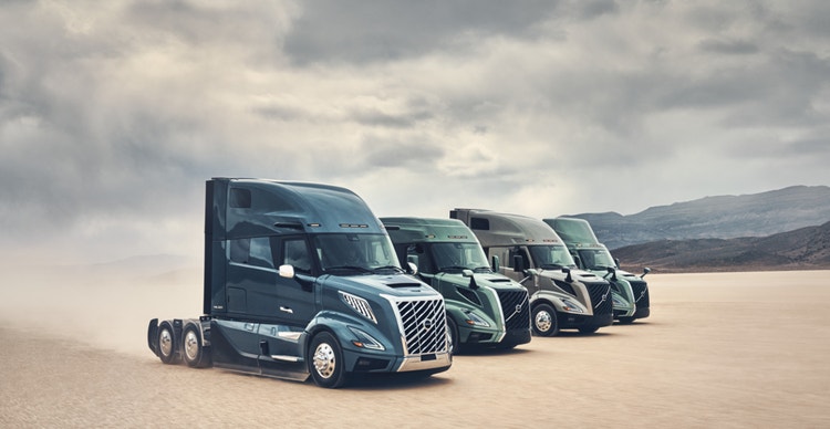 A lineup of four Volvo VNL trucks in colors Coastal Mist, Prairie Grasslands, Desert Mirage and Skyline Steel, moving across a vast desert plain with mountains in the distance.