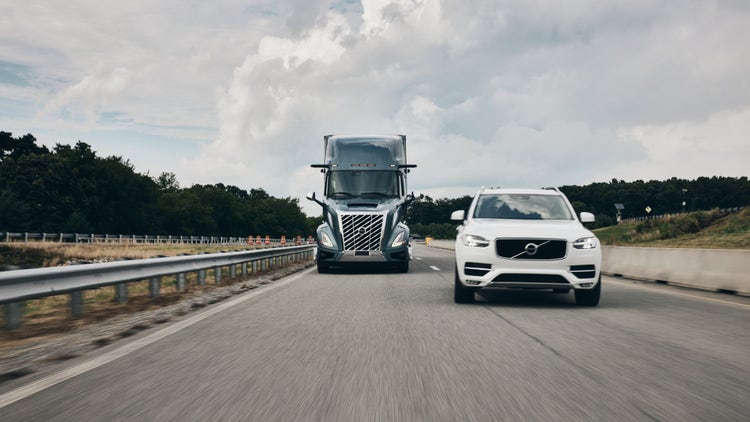 A Volvo VNL truck and a white Volvo SUV driving side by side on a highway, with trees lining the road.