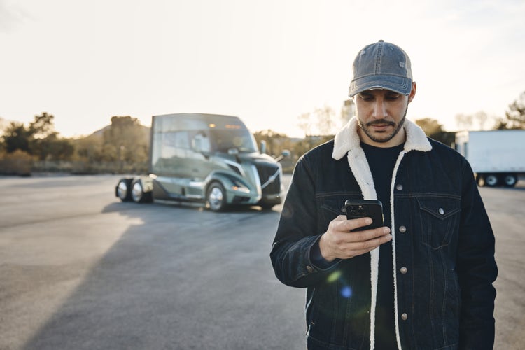 A man wearing a denim jacket and cap looks at his smartphone in a parking lot, with a Volvo VNL truck parked in the background.