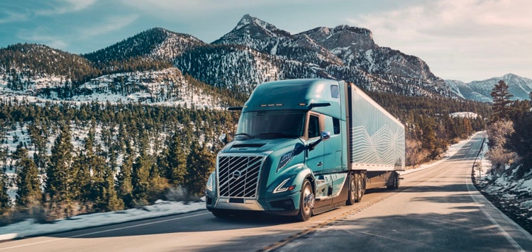A Coastal Mist Volvo VNL truck hauling a trailer on a scenic mountain road, surrounded by snow-dusted trees and peaks under a clear sky.