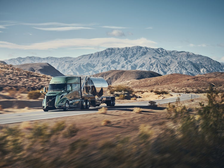 A Volvo VNL truck hauling a silver tanker trailer on a winding desert road with mountains in the background.
