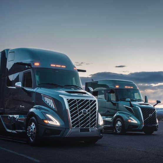 Two Volvo VNL trucks parked side by side at dusk, their LED lights illuminated against a backdrop of a darkening sky.