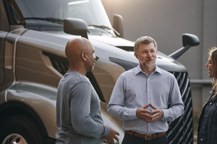 Three people stand and converse near the front of a parked Volvo VNL truck, with its grille and side mirror visible in the background.