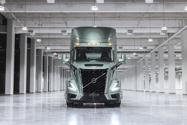 A front-facing Volvo VNL truck displayed in a bright, spacious indoor setting with rows of overhead lights and white structural columns.