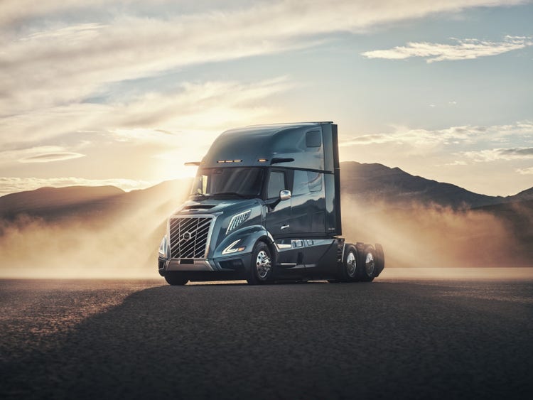 A Volvo VNL truck driving through a dusty open area, with low hills in the background