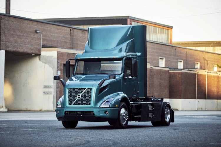 Volvo VNR Electric truck parked in an industrial area, with a teal cab, aerodynamic design, and clean lines, against a backdrop of brick buildings.