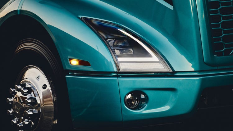 Front end of a Volvo VNR Electric truck in teal, showcasing its sleek headlight design, side marker light, and polished chrome wheel.