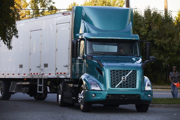A teal Volvo VNR Electric truck with a box trailer stopped on a street.