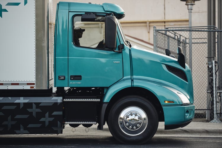 Side profile of a teal Volvo VNR Electric truck parked next to a fence. The sleek design showcases the cab, front wheel, and aerodynamic features.