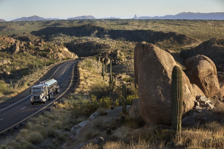 A Volvo VNR tanker truck driving on a winding road through a scenic desert landscape