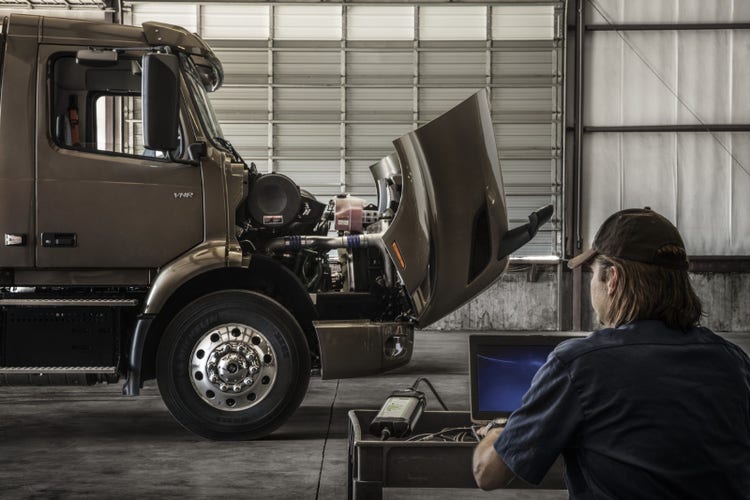 A mechanic using diagnostic equipment connected to a laptop while inspecting a Volvo VNR truck with its hood open in a service garage.