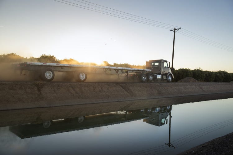 A Volvo VNR truck with a flatbed trailer driving alongside an irrigation canal at sunset, kicking up dust in the rural landscape.
