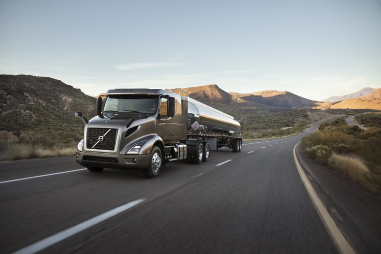 A Volvo VNR truck hauling a polished tanker trailer on a winding highway through a desert landscape