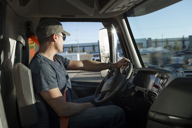 A truck driver in a Volvo VNR cab glances out the window, seated in a modern interior with a visible dashboard and orange seatbelt.
