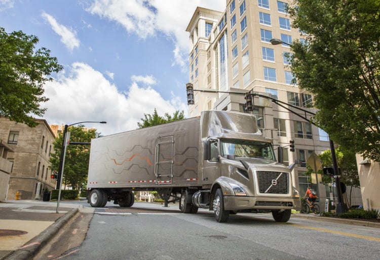 A Volvo VNR truck with a box trailer navigates a city intersection, surrounded by modern buildings and trees.
