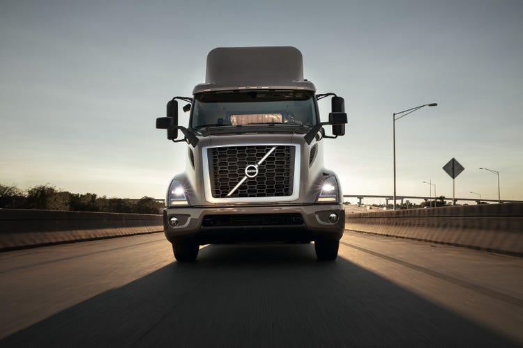 Front view of a Volvo VNR truck driving on a highway at sunrise, with its headlights illuminated and a clear sky in the background.