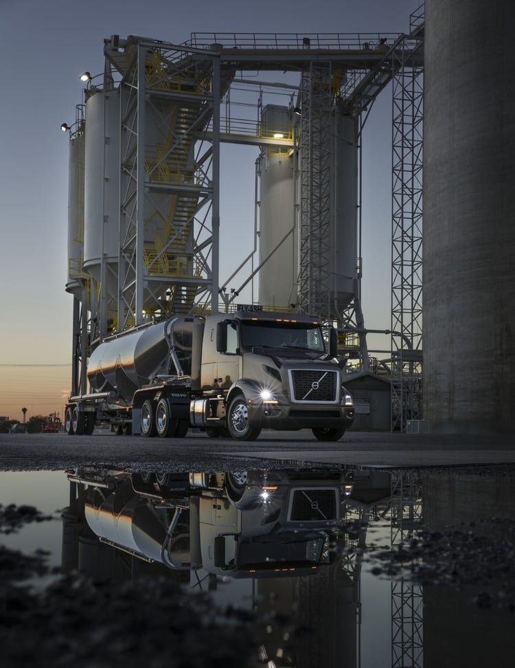 A Volvo VNR truck with a tanker trailer parked at an industrial facility, reflected in a puddle at dusk.