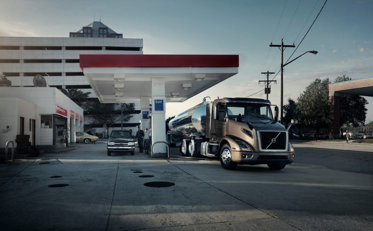 A Volvo VNR truck with a polished tanker trailer parked at a gas station, alongside a pickup truck and other vehicles under a clear sky.