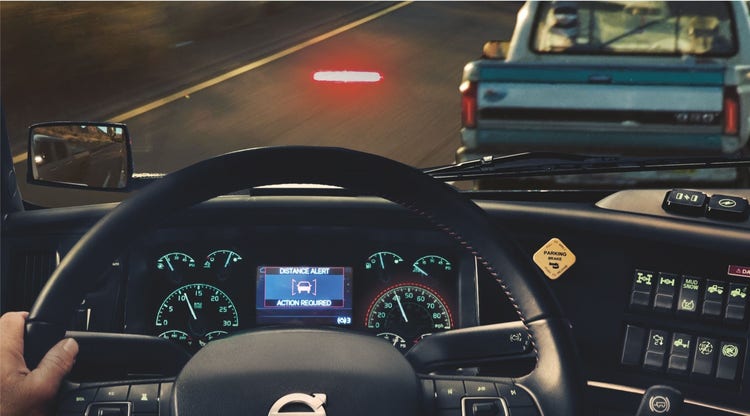 View from the driver’s seat of a Volvo truck showing the dashboard and a "Distance Alert" warning on the digital display as the truck approaches another vehicle on the road.