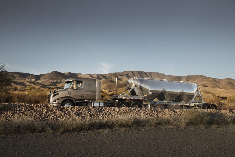 Side view of a Volvo VNR truck pulling a shiny tanker trailer along a gravel road, set against a backdrop of rugged desert hills.