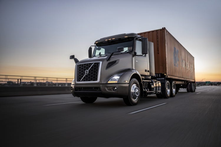 A Volvo VNR truck hauling a shipping container on a highway at dusk