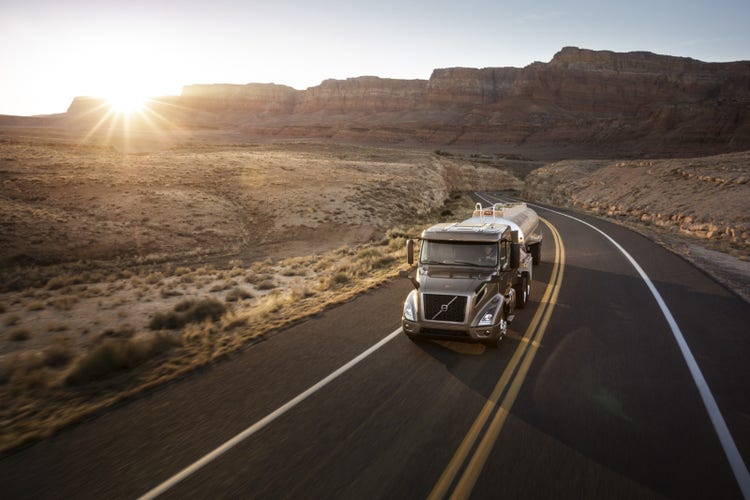 A Volvo VNR truck hauling a tanker trailer on a winding desert highway at sunrise, with red rock cliffs in the background.