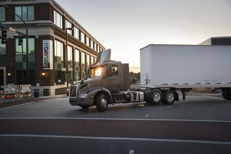 A Volvo VNR truck with a box trailer stops at an urban intersection, framed by a modern brick building and road closure signs.