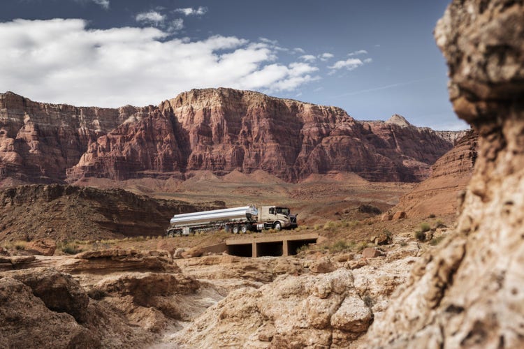 A Volvo VNR truck hauling a polished tanker trailer through a rugged desert canyon, with towering red rock cliffs in the background.