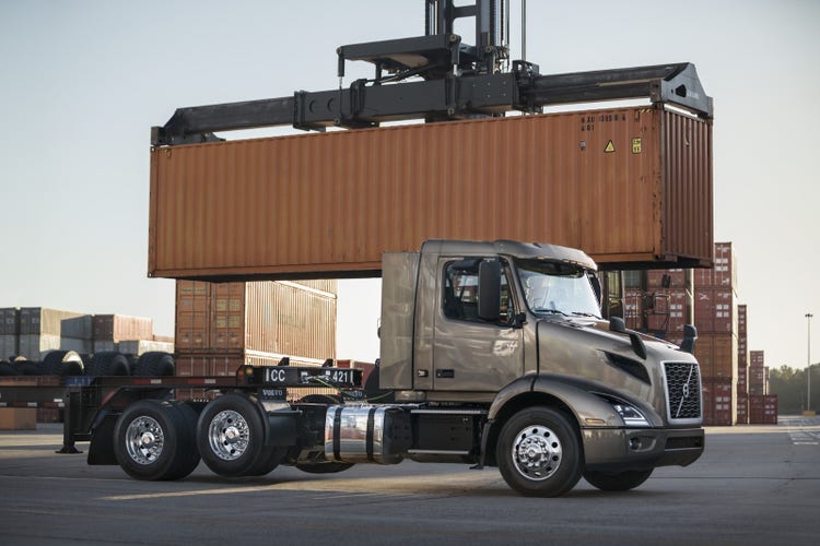 A Volvo VNR truck parked in a shipping yard, positioned near a crane lifting a large orange cargo container.